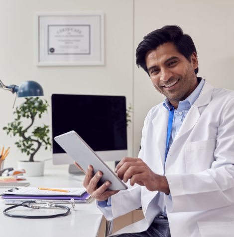 Portrait Of Male Doctor Or GP Wearing White Coat Sitting At Desk In Office Using Digital Tablet
